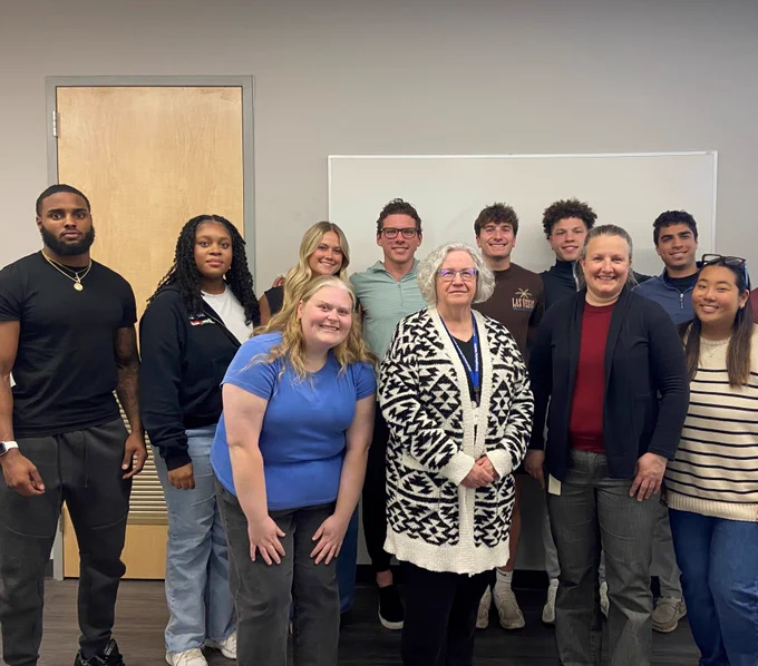 A group of eleven people standing closely together indoors in front of a whiteboard. They are diverse in age, race and gender. One woman in the front is wearing a black and white cardigan.