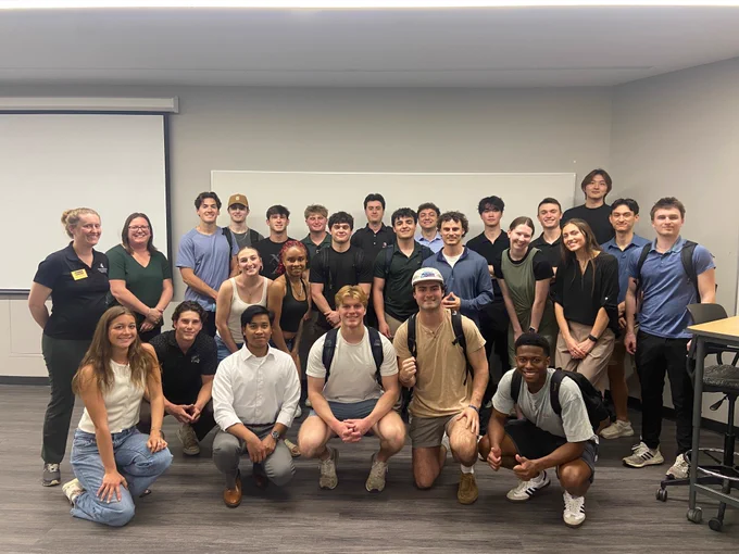 Group photo of about 30 students and 2 staff in a classroom, posing in rows. Some students are squatting in the front row. A whiteboard and projector screen are in the background.