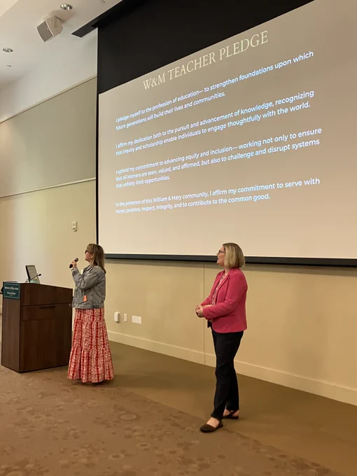 Two people stand at the front of a university auditorium during a formal event. One speaks into a microphone at a podium while the other stands nearby. A large projected slide behind them reads ‘W&M Teacher Pledge,’ setting a reflective, ceremonial tone for the program.