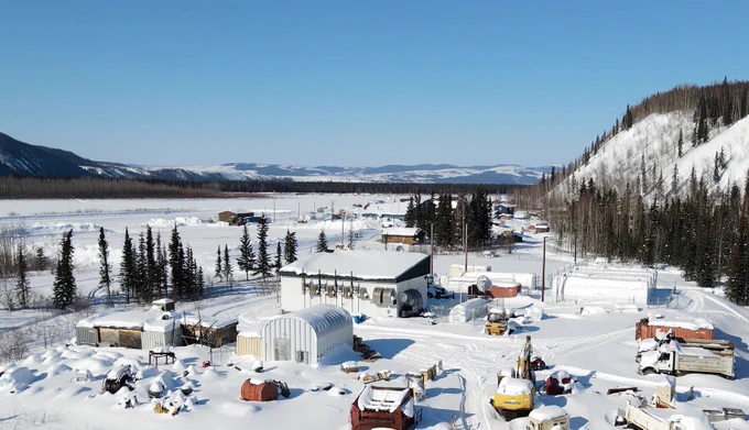 A snowy, remote area in a mountainous region. The landscape includes several buildings and storage tanks, with evergreen trees scattered around. Snow-covered roads lead through the facility, and distant hills are visible under a clear blue sky.
