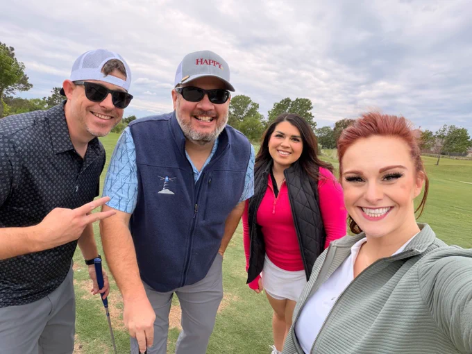 Group of four friends posing on a golf course with cloudy sky in the background, dressed in casual golf attire.