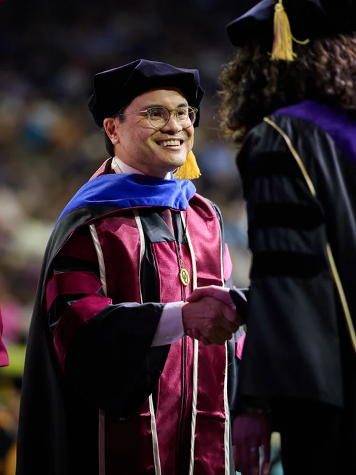 A person in graduation attire, including a maroon and black gown with a blue hood, shakes hands with another similarly dressed individual. The setting is a graduation ceremony.