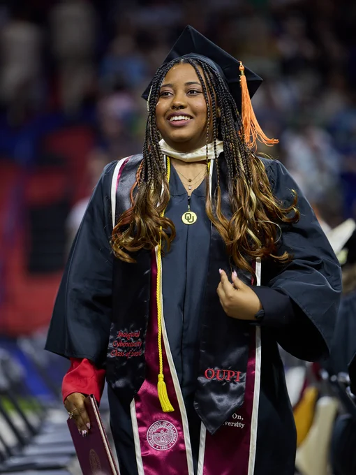 This image shows a graduate in a cap and gown holding a diploma. The gown features the letters "OUPI." The scene is a graduation ceremony in progress, with other graduates and attendees blurred in the background.