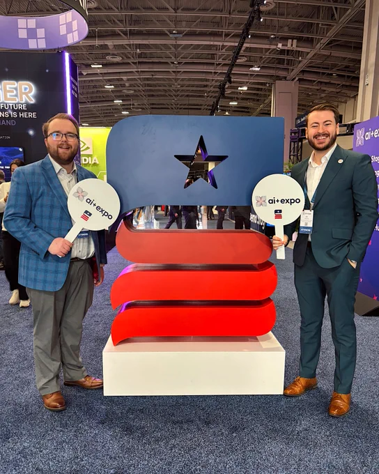 Two individuals in suits are smiling and standing in front of a large sculpture of waves and a star, reminiscent of a flag, at the AI Expo event. They are holding circular signs that read "ai expo." The setting appears to be an indoor exhibition hall.