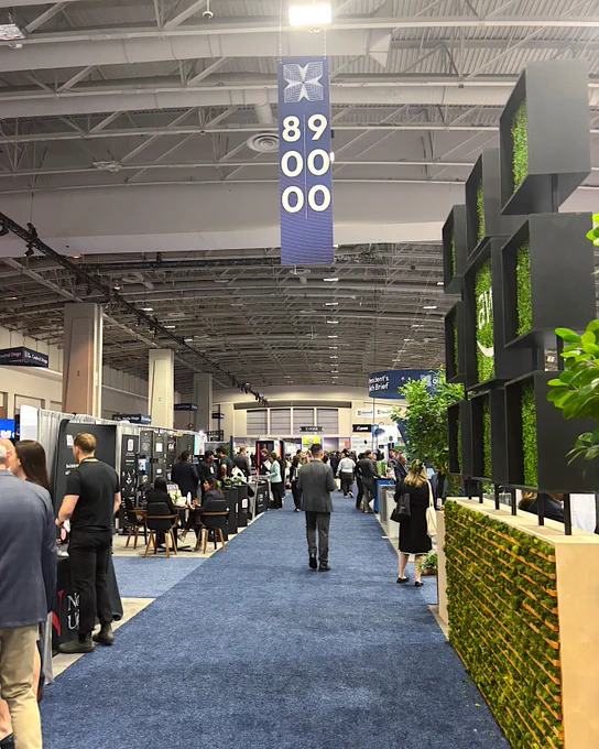 A spacious exhibition hall with a blue carpeted aisle is seen. There are several booths on both sides featuring displays and people engaged in conversation. A sign above indicates the area as "89000." Prominent greenery decorates the right side, adding a touch of nature to the scene.