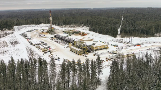 Aerial view of a snowy landscape featuring an oil drilling site surrounded by dense forest. The site includes various industrial structures, vehicles, and equipment. Snow covers the ground and trees.
