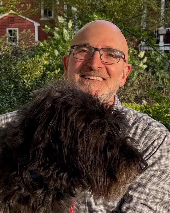 A smiling person (Daniel Promislow) with glasses holds a shaggy black dog in front of a blurred background of greenery and a red building.