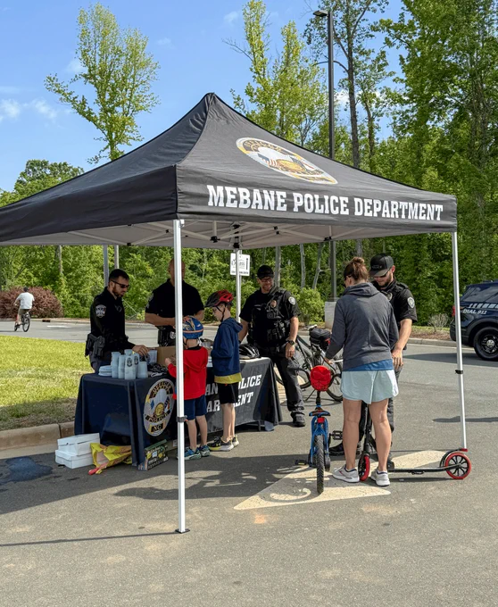 Mebane Police Department officers engaging with children and adults under a black canopy tent during a community event.