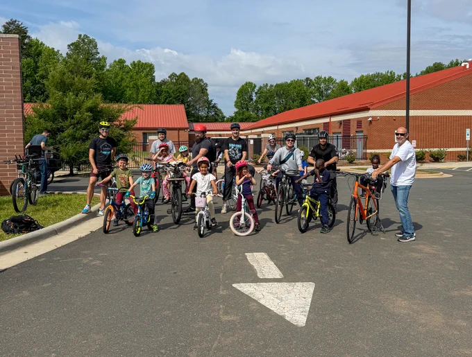 Group of adults and children posing with bicycles on a sunny day in a school parking lot.
