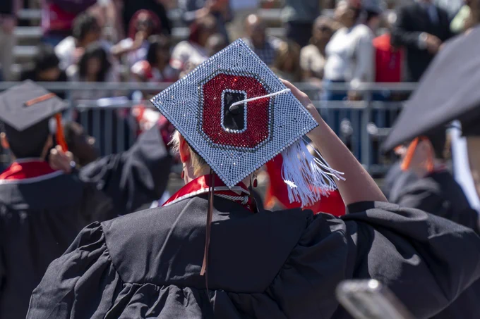 The view from behind an Ohio State graduate, their cap is fully bedazzled with a scarlet Block O