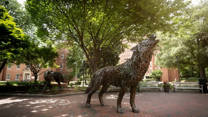 Metal sculpture of a howling wolf standing on a brick pathway in NC State's Wolf Plaza, surrounded by trees.