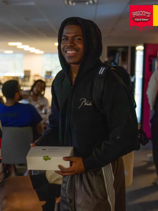 A young man with a backpack smiles broadly while holding a box at an indoor event. The setting appears social, with other attendees visible in the background.