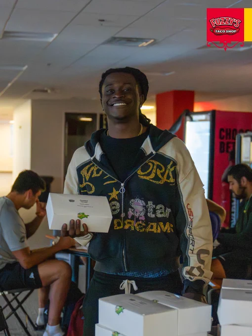 A person smiling and holding takeout boxes from Fuzzy's Taco Shop at a social gathering. The mood is cheerful and relaxed.