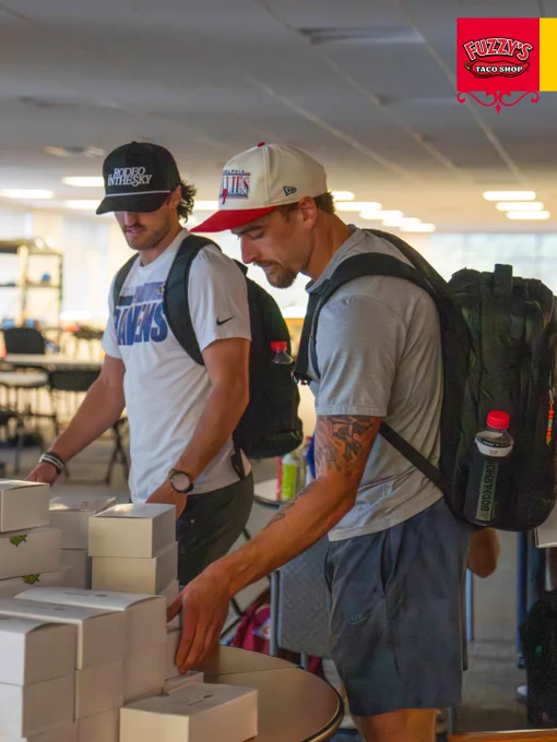 Two individuals are picking up boxes from a table at a gathering, creating a casual and laid-back atmosphere. They appear to be in a social setting, with backpacks and casual attire.