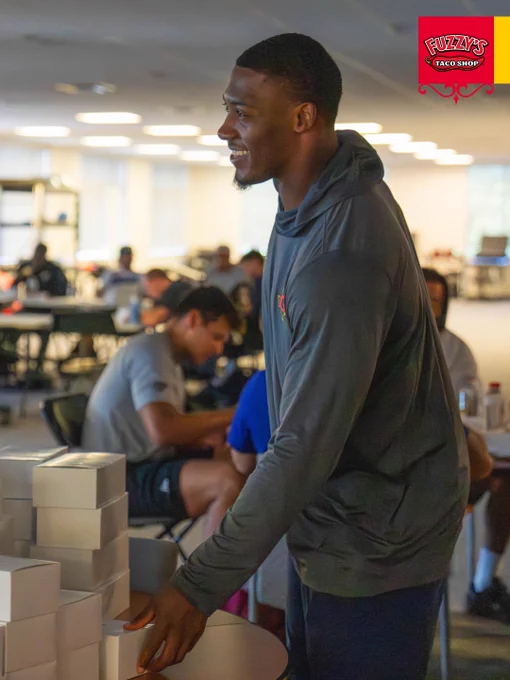 A smiling man stands indoors near a stack of white boxes, suggesting organization or event preparation in a casual setting.