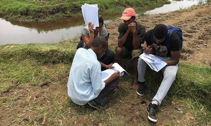 Four people are sitting on grassy ground near a small body of water, examining and discussing papers. One person is partially covering their face with papers. The scene suggests a group outdoor study or project discussion.