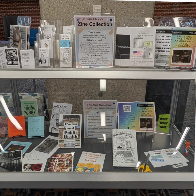 A glass and metal library display case with books and zines in the case. There are zines on the top of the case.