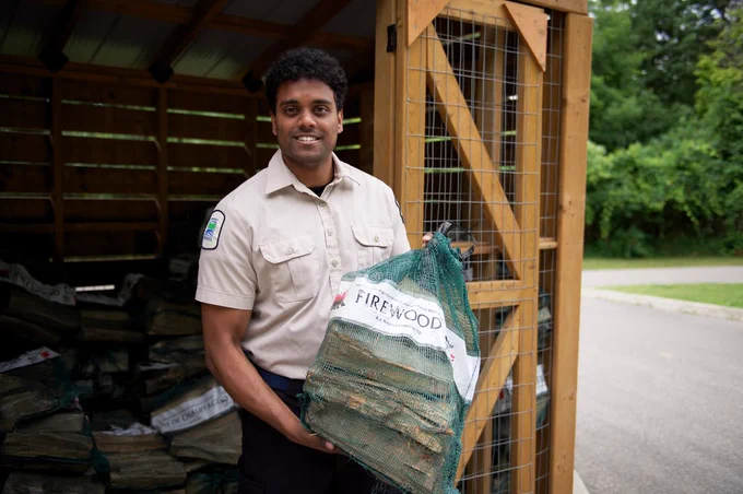 A person in a uniform stands smiling, holding a net bag of firewood. 