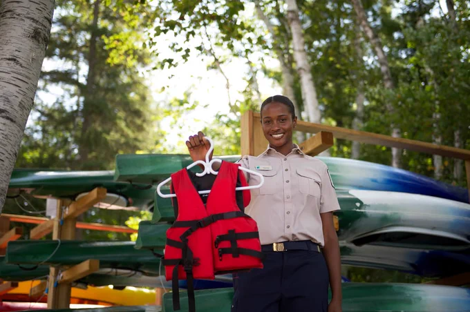 A park ranger in uniform is standing outdoors, holding a red life jacket on a hanger. 