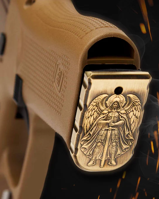 Close-up of a brass St. Michael engraved magazine plate installed on a tan pistol grip, surrounded by subtle smoke and glowing ember effects against a dark background.