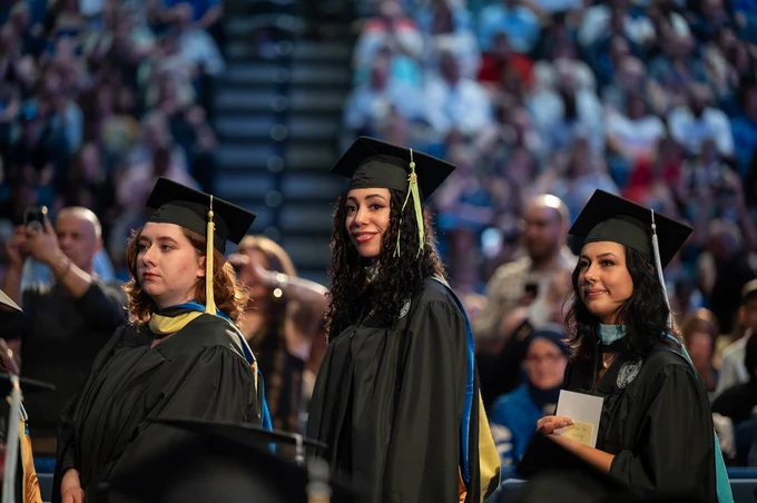 Graduation ceremony: Three grads in caps and gowns stand smiling. Center grad wears a cap with '25' and curly hair. Crowd blurs behind. 