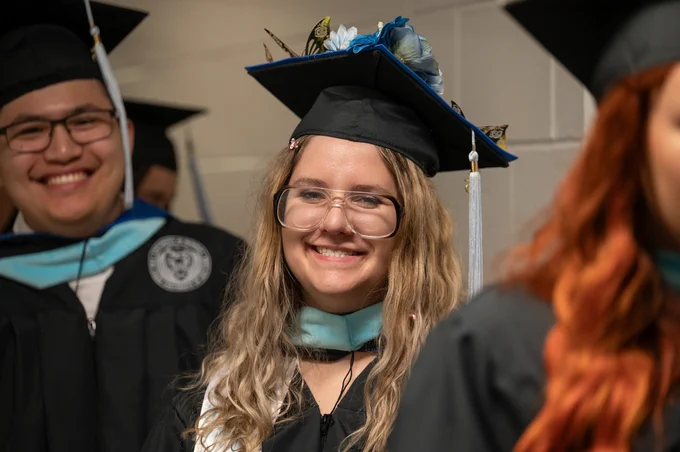 Grad with wavy blonde hair and glasses smiles and has flowers on cap. 