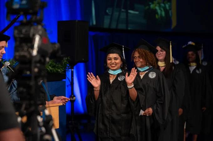 Graduates in black gowns & caps walk during commencement. A smiling grad waves, wearing a light blue mask, followed by others. 