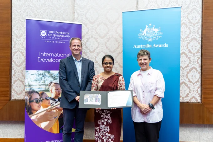A participant with both course leads stand indoors holding a framed certificate, flanked by banners for The University of Queensland and Australia Awards programs.