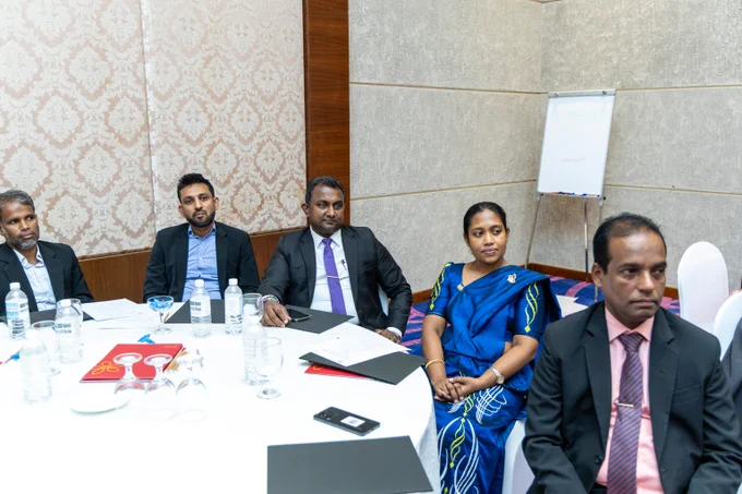Five participants seated around a round table in a conference room with documents and water bottles in front of them.