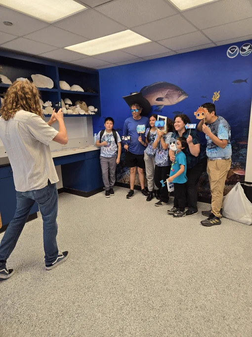 A group of seven poses for a photo in front of a wall with a large photo of coral and fish on it. Another person takes their photo.