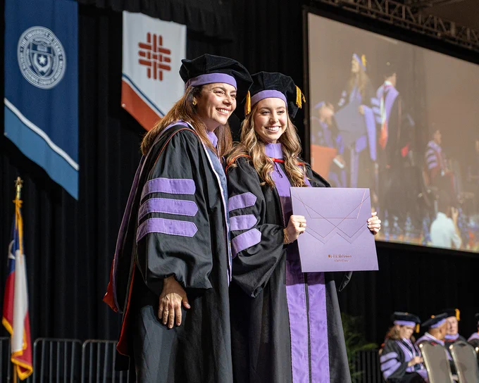 A faculty member in black doctoral regalia with purple velvet panels stands on stage beside a graduate who is holding a large purple diploma cover labeled “UTHealth Houston School of Dentistry.” Behind them hang banners, including one with the UTHealth Houston seal and another with the university’s orange tapestry, while a large screen shows other graduates in similar attire crossing the stage.