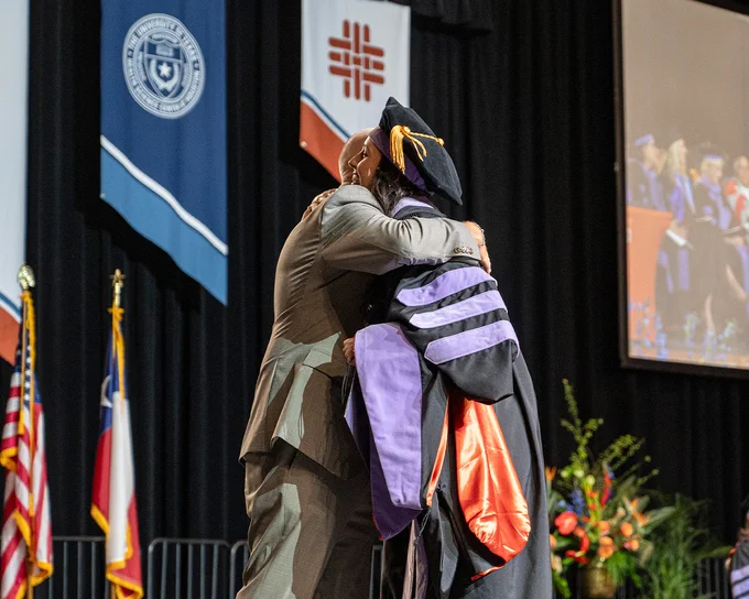 A graduate in black doctoral regalia with purple and orange accents hugs a family member on stage during a commencement ceremony. Behind them are university banners, the Texas and U.S. flags, and a large screen showing a live feed of the event.