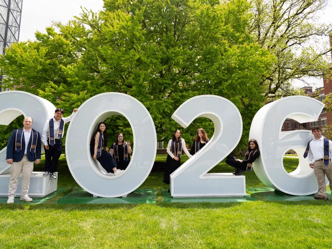 A group of students in commencement attire pose for a photo with giant letters that spell out UR2026. 