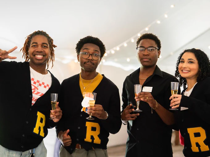 A group of four students wearing cardigans with the letter R pose for a photo holding up a glass of champagne.