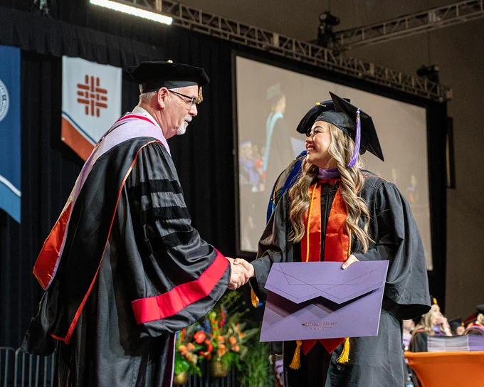 A graduate in a black cap and gown with an orange stole and purple hood shakes hands with a faculty member in black academic regalia with red accents and a purple hood. The graduate holds a large purple diploma cover labeled “UTHealth Houston School of Dentistry.” Behind them are hanging banners, floral arrangements, and a large screen displaying a stream of the commencement ceremony.