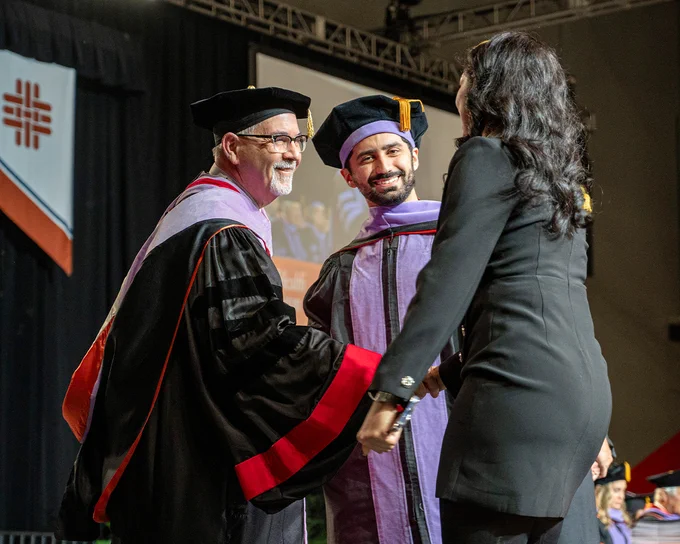 A faculty member in black academic regalia shakes hands with a woman accompanying a male graduate across the stage during a commencement ceremony. Behind them are university banners, the Texas and U.S. flags, and a large screen showing a live feed of the event.