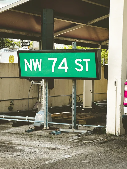 A green road sign that reads 'NW 74 ST' in bold white letters, mounted on a weathered metal structure. Overhead is a carport-like structure. A Miami-Dade Corrections vehicle is visible in the background, along with 'ONE WAY' sign.