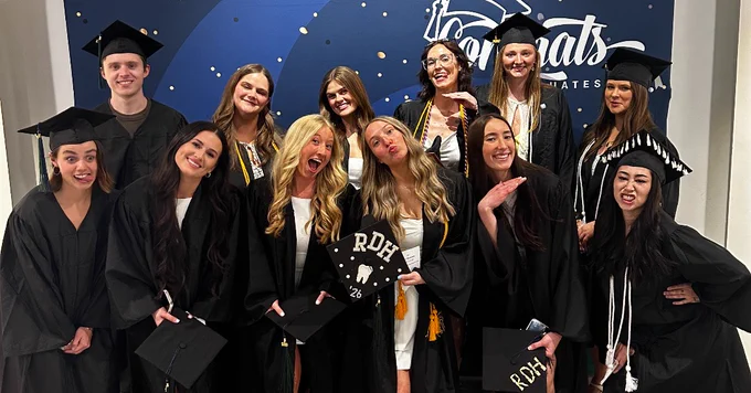 A group of people in graduation attire stand in front of a backdrop that says "Congrats Grad." They are wearing caps and gowns, and some are holding diplomas. The setting appears to be indoors.