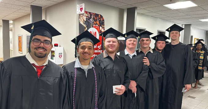 A group of people in graduation gowns and caps stand in a hallway, smiling at the camera.