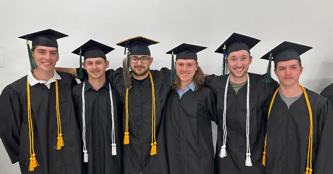 A group of six individuals wearing graduation caps and gowns stands in front of a plain wall. They are smiling, with some having their arms around each other.