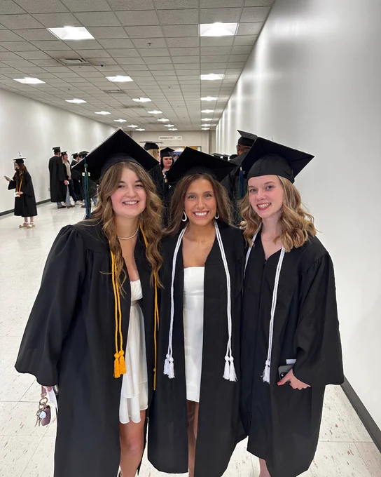 Three graduates smile for a photo. They are wearing black caps and gowns with various colored cords around their necks.