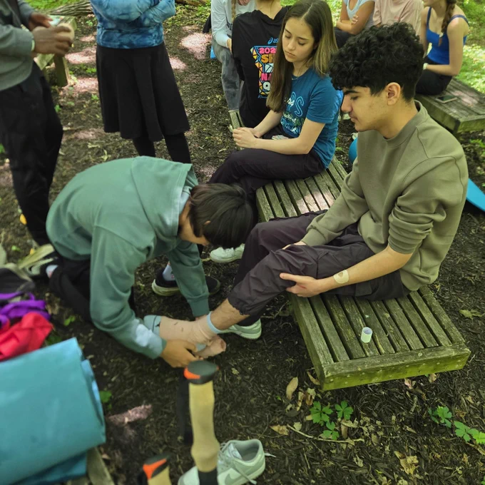 Several people sitting on a wooden bench in a wooded area, interacting with one another.