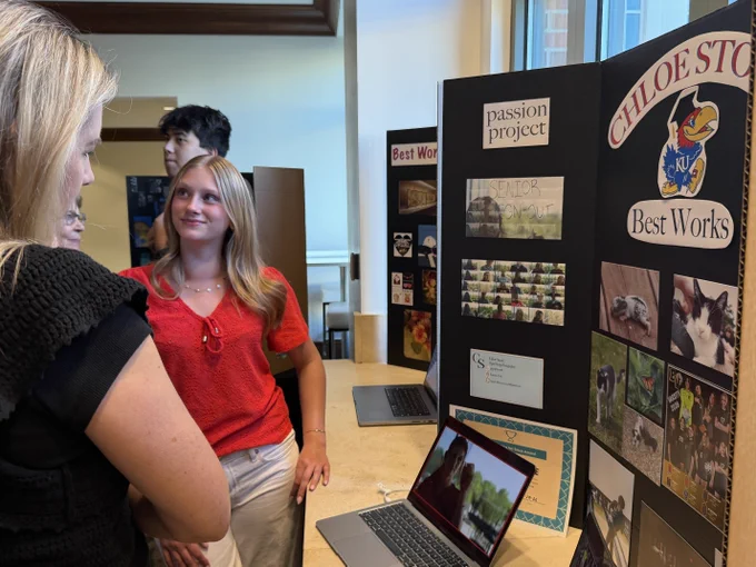 Two women standing in front of a display board showing images, graphic & photography, and text at the CAPS showcase.