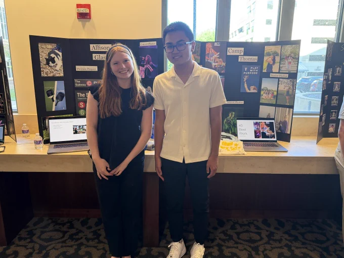 Two young adults, a woman and a man, standing together in front of a display board with various media and graphic design images at the CAPS showcase.