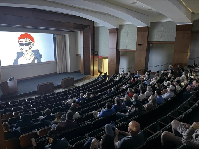 A movie theater interior with a large projection screen and rows of people seated in the audience.