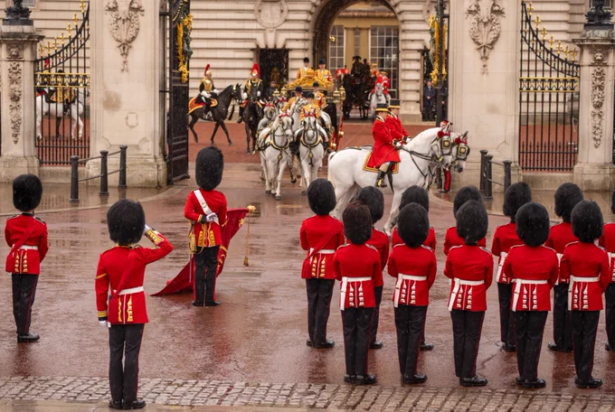 A group of ceremonial guards in red uniforms with tall black hats stands in formation outside Buckingham Palace. A horse-drawn carriage escorted by mounted guards passes through the palace gates. The setting is wet from rain.