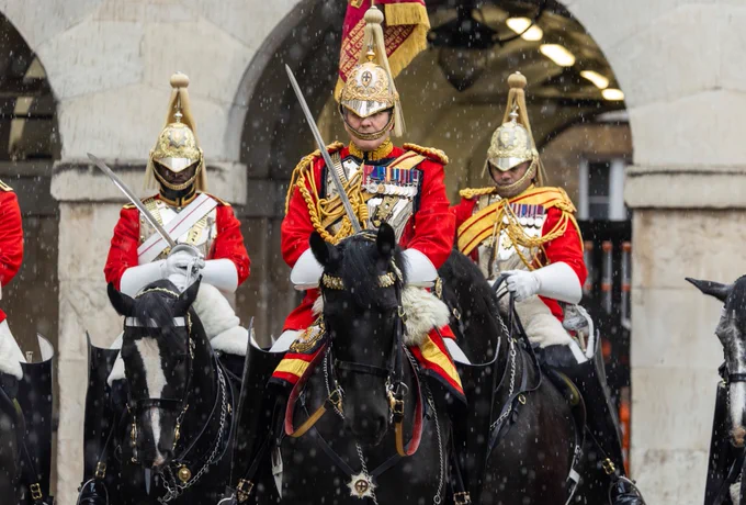 Members of the Household Cavalry in ceremonial uniform, including gold helmets and red tunics, riding black horses in an official procession. Snow is gently falling, and stone arches are visible in the background.