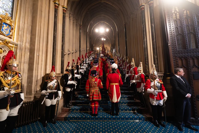 This image shows a ceremonial procession inside the entrance of Westminster Palace. Figures in traditional red and gold military uniforms stand on a grand staircase under an ornate arched ceiling.