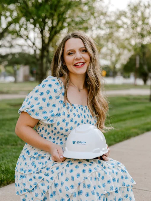 A young lady posing by squatting down in a sitting position holding a white hat labeled Valero. The lady has dirty blonde hair and is wearing a white sun dress with blue flowers. 