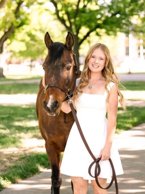 A young lady with blonde hair wearing a white dress standing next to a brown horse holding the reigns. The background is filled with green leaves and sunshine. 